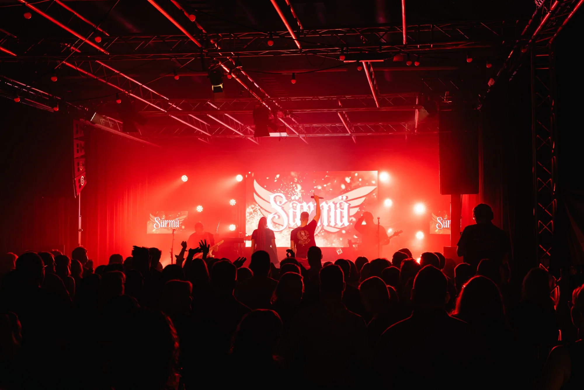 Yhtyeen konsertti punaisessa valossa, yleisön siluetit ja lavan taustanäytöllä siivekäs logo