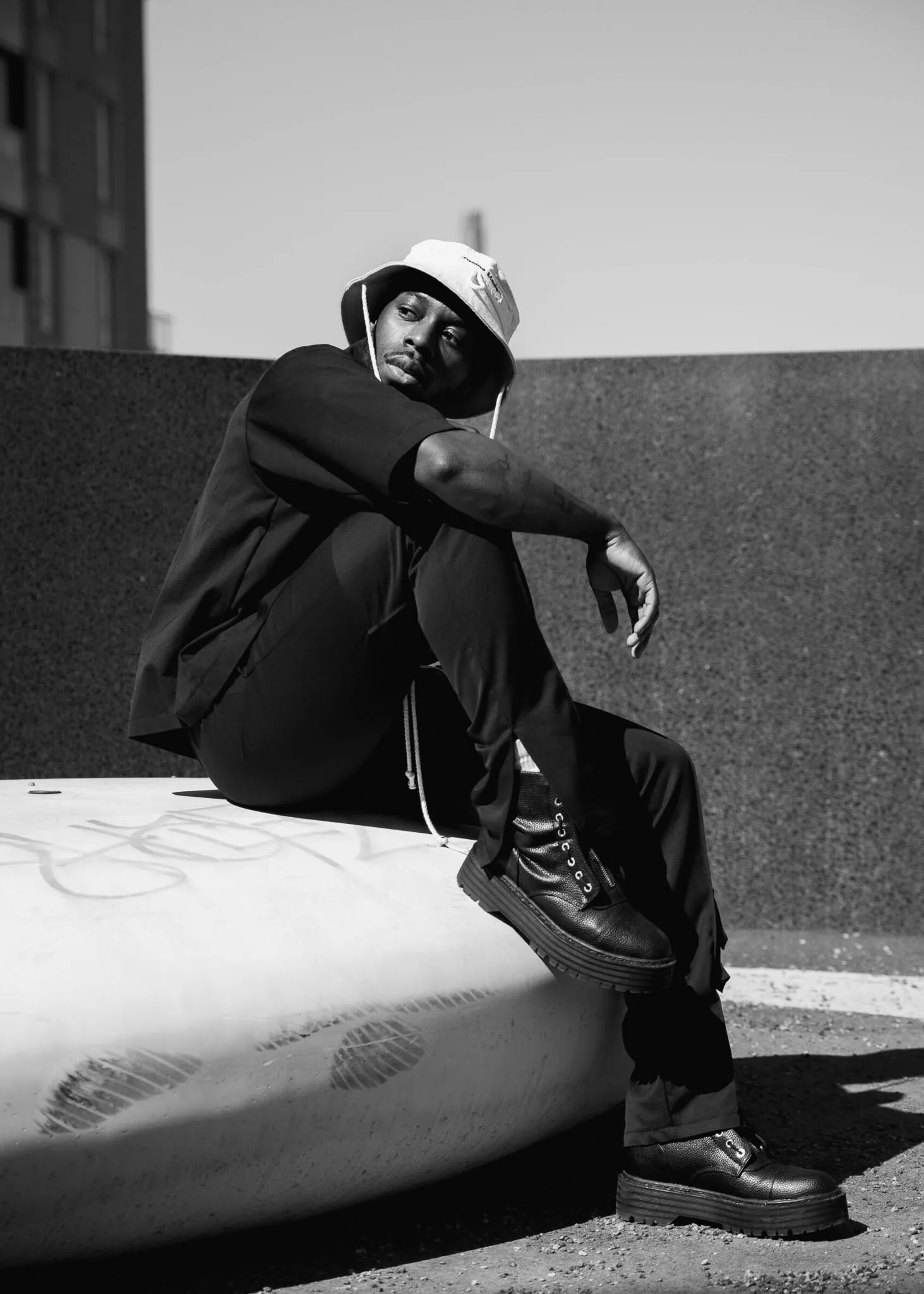 Mustavalkoinen muotokuva: streetwear-asuinen henkilö bucket-hatussa istumassa betonilohkolla