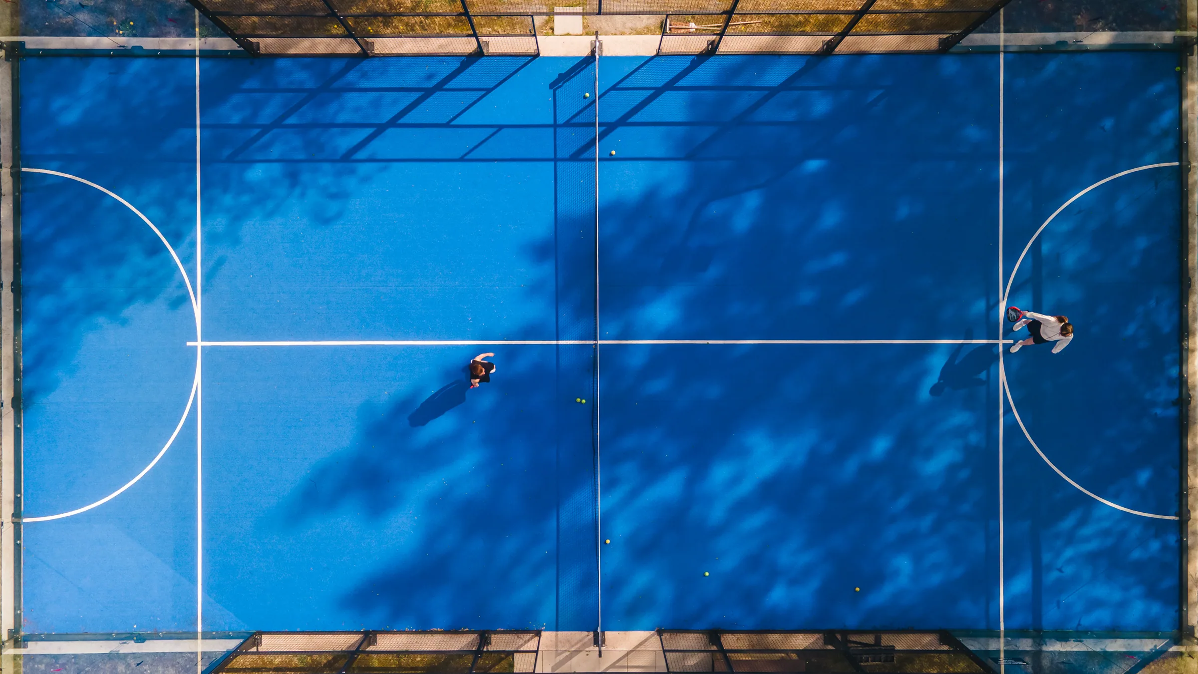 Ilmakuva suoraan ylhäältä sinisestä padel-kentästä ja pelaajista
