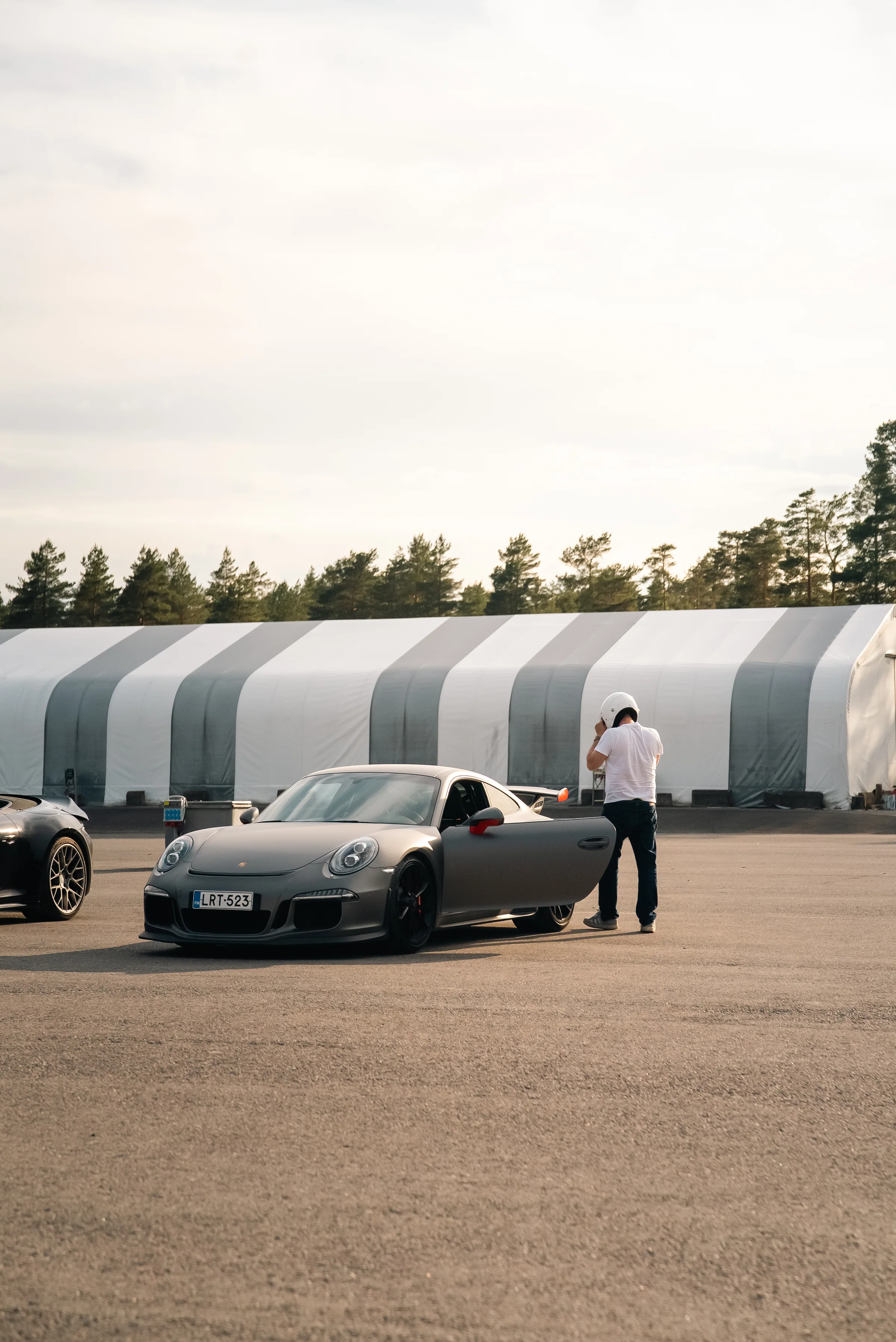 Kuljettaja pukee kypärää mattaharmaan Porsche 911 GT3:n vieressä varikolla, taustalla halliteltat, ratapäivä Alastarolla